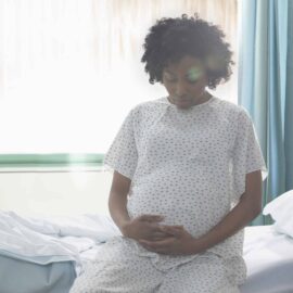 A Black pregnant woman holds her belly as she sits on a bed in a hospital.